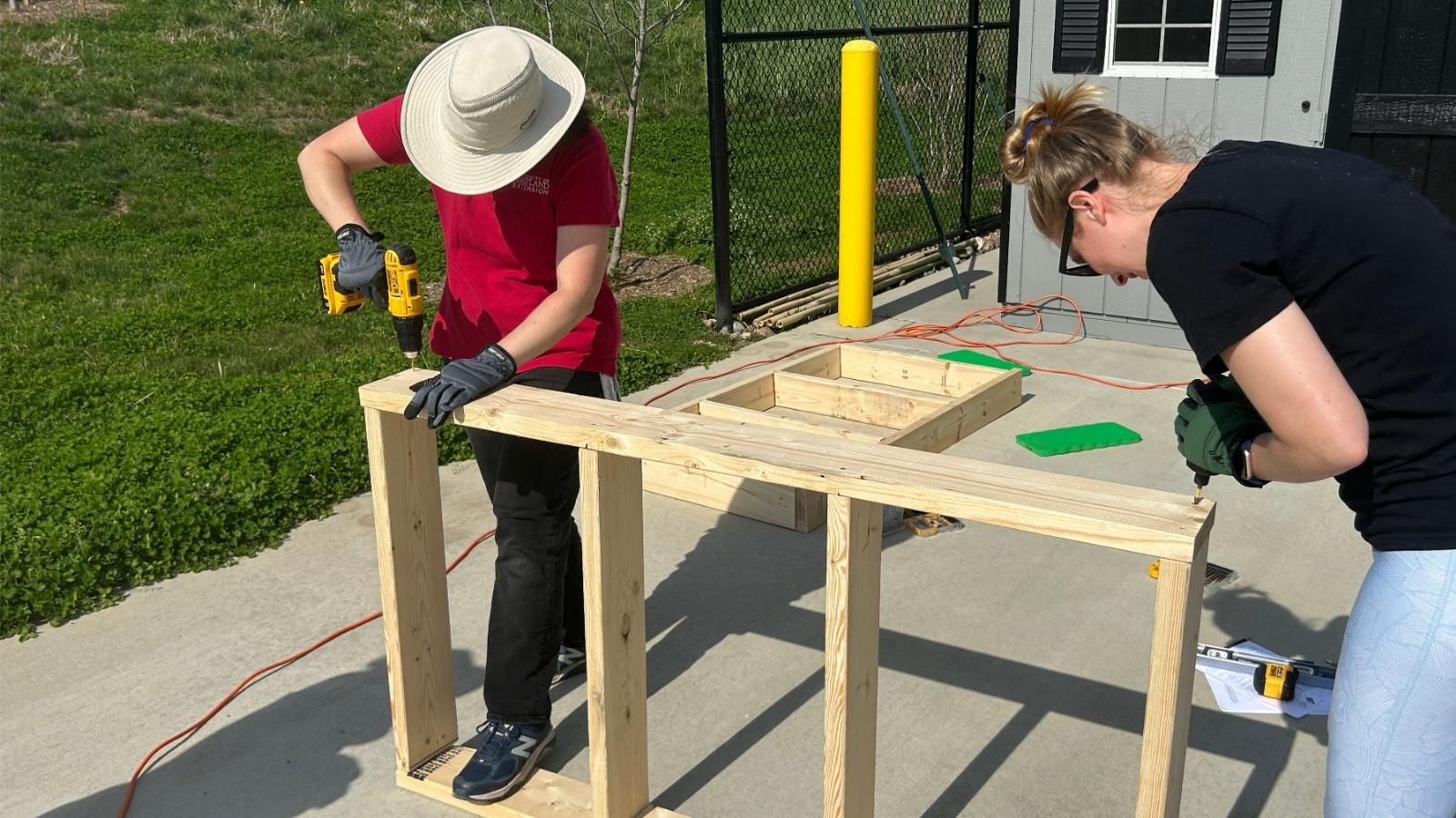 Two people use cordless drills to screw together pieces of a wooden table frame.