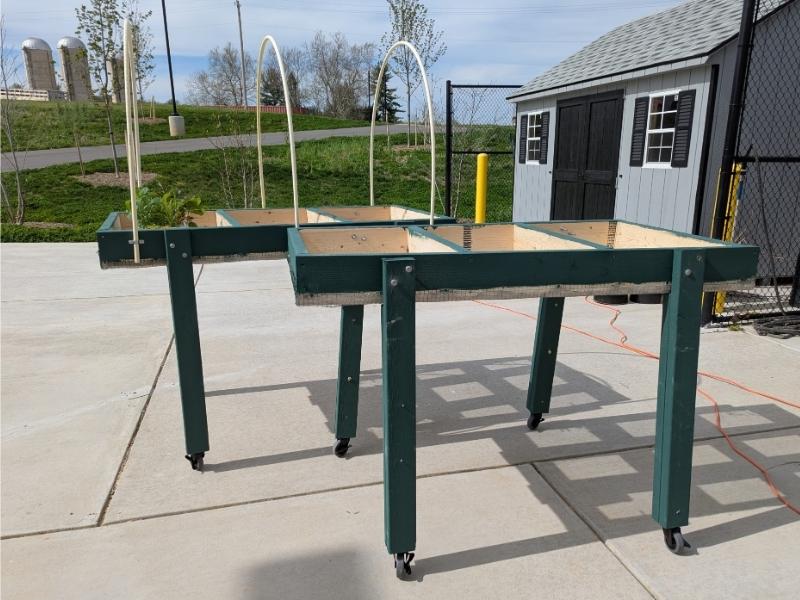 Two empty, fully-built Salad Tables, painted on the exterior wood surfaces. One table has 3 pieces of slender PVC pipe arched over the table, anchored with brackets on the table's sides.