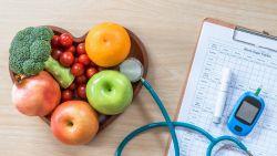 heart shaped wooden bowl filled with fruit and vegetables next to a stethescope and glucose monitor and a blood sugar tracker