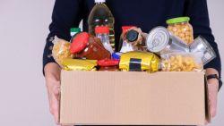 brown box filled with canned goods and pantry food held by a white man wearing a black watch and a dark blue sweater, only his torso is shown