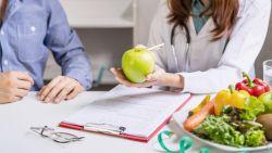a woman holding an apple over a clipboard, seemingly talking to a man sitting next to her, near the clipboard there are glasses and a plate of vegetables