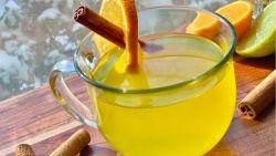 Glass mug filled with citrus turneric tea with a cinnamon stick coming out on top of a wooden cutting board