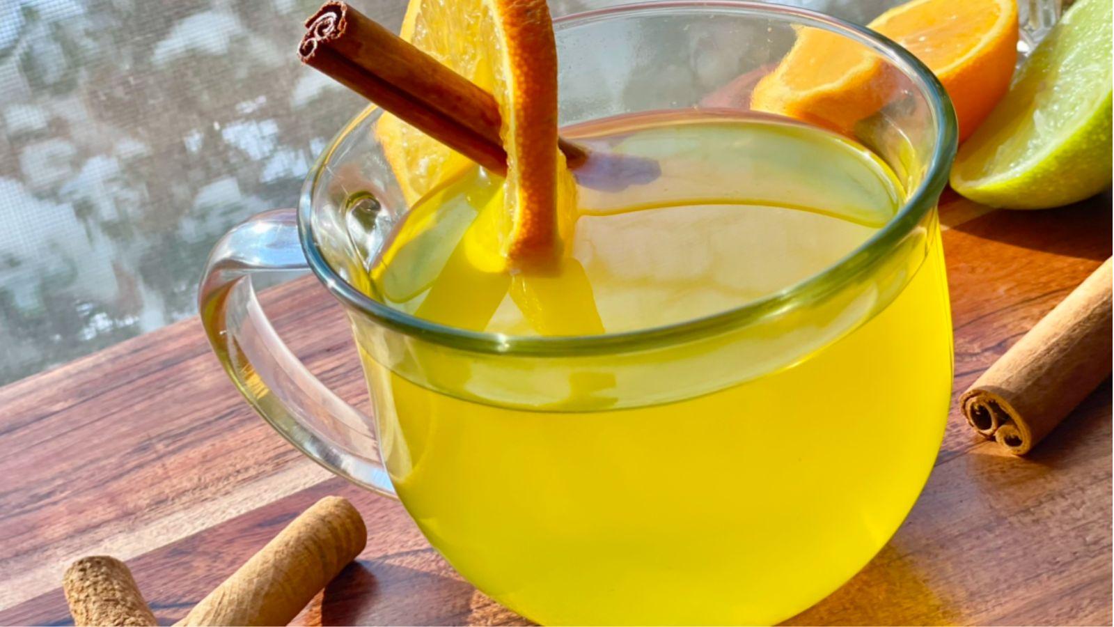 Glass mug filled with citrus turneric tea with a cinnamon stick coming out on top of a wooden cutting board