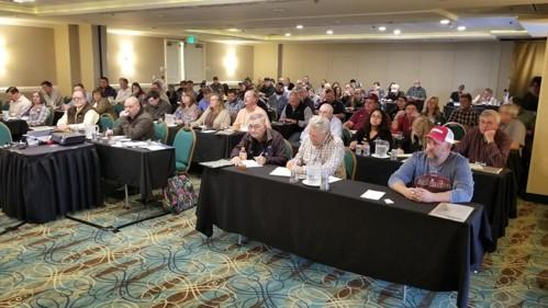 Attendees seated at tables during an agricultural education meeting or conference, facing a presenter outside the image. The room is arranged classroom-style with participants taking notes and listening to the presentation.